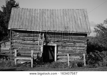 This black and white image of a sawn log cabin with a door and tin roof show the long side of the building.