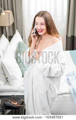 Portrait of smiling woman in the white robe with mobile phone in the cozy hotel room or bedroom