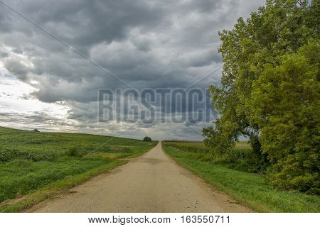 A straight and narrow rural road stetches to the horizon amidst green fields beneath a summer sky threatening rain.