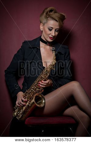 Sexy attractive woman with saxophone posing on red background. Young sensual blonde playing sax. Musical instrument, jazz. Beautiful woman in black posing with saxophone, studio shot