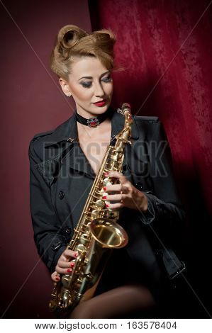 Sexy attractive woman with saxophone posing on red background. Young sensual blonde playing sax. Musical instrument, jazz. Beautiful woman in black posing with saxophone, studio shot