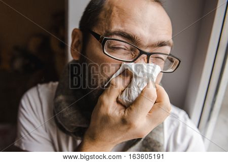 Young man blowing his nose while sitting at the window