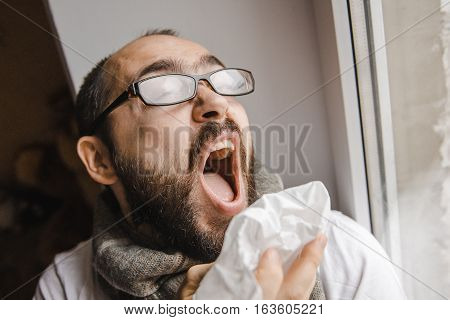 Young man blowing his nose while sitting at the window