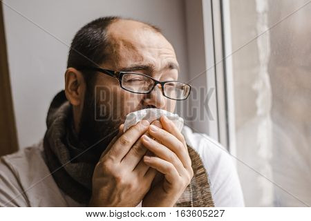 Young man blowing his nose while sitting at the window