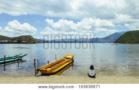 Single young Chinese woman looks into skyline, sitting on bank of Lugu lake, Lijiang, China, back to camera