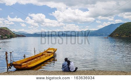 Young Chinese couple sits on beach, leaning on each other, looking to skyline of Lugu Lake, at Xiaoluoshui village, Lijiang, Yunnan, China