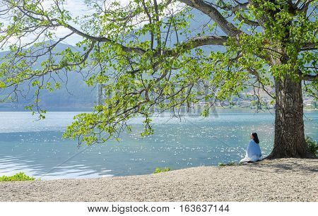 Single young Chinese woman looks up, sitting under big tree on bank of Lugu lake, back to camera,  at Daluoshui village, Lijiang, China