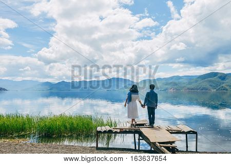 Young Chinese couple hold each other, standing on wasted wooden platform hand in hand on bank of Lugu lake, back to camera,  at Daluoshui village, Lijiang, China