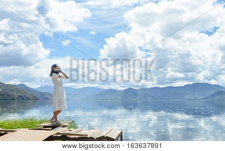 Single young Chinese woman in white long skirt stands on wasted platform on bank of Lugu lake, listening to music, back to camera, at Daluoshui village, Lijiang, China
