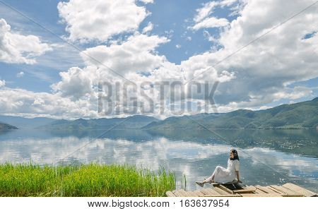 Single young Chinese woman in white long skirt sits on wasted platform on bank of Lugu lake, listening to music, back to camera, at Daluoshui village, Lijiang, China