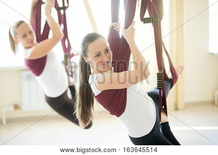 Two young attractive happy smiling yogi women doing aerial yoga practice in purple hammocks in fitness club. Beautiful females working out in class, performing antigravity yoga