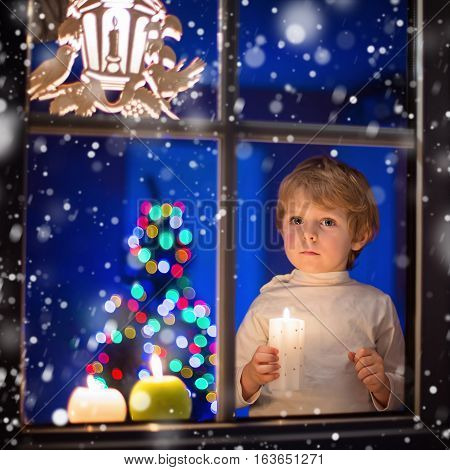 Beautiful little kid boy, standing by window at Christmas time and holding candle. Child waitng for Santa. With colorful lights from Christmas tree on background.