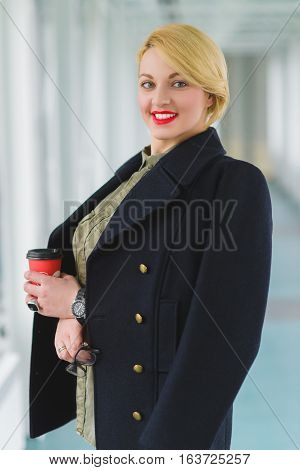 Portrait of blond businesswoman holding coffee and glass in lobby.