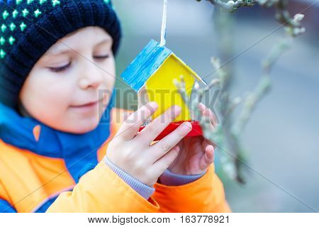 Little kid feeding birds in winter. Child hanging colorful selfmade bird house on tree on frosty cold day. Toddler in colorful wam clothes. Selective focus on hands and feeder with seeds