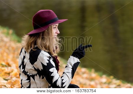 Woman in a floral patterned coat and wine red hat in the park by the river. Happy girl and colorful autumn forest. Portrait of a lady yellow green red and brown leaves background
