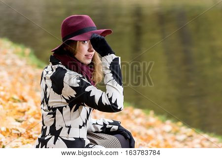 Woman in a floral patterned coat and wine red hat in the park by the river. Happy girl and colorful autumn forest. Portrait of a lady yellow green red and brown leaves background
