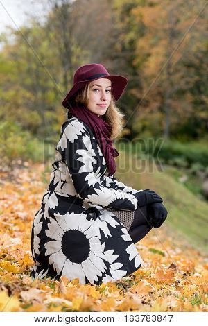 Woman in a floral patterned coat and wine red hat in the park. Happy girl and colorful autumn forest. Portrait of a lady yellow green red and brown leaves background