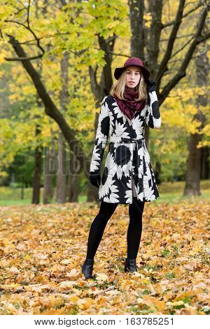 Woman in a floral patterned coat and wine red hat in the park. Happy girl and colorful autumn forest. Portrait of a lady yellow green red and brown leaves background