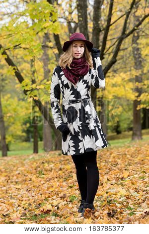 Woman in a floral patterned coat and wine red hat in the park. Happy girl and colorful autumn forest. Portrait of a lady yellow green red and brown leaves background