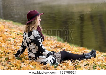 Woman in a floral patterned coat and wine red hat in the park by the river. Happy girl and colorful autumn forest. Portrait of a lady yellow green red and brown leaves background