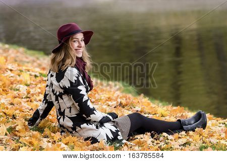 Woman in a floral patterned coat and wine red hat in the park by the river. Happy girl and colorful autumn forest. Portrait of a lady yellow green red and brown leaves background