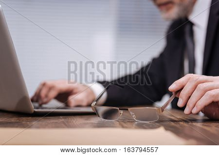 I need these. Neat well dressed qualified office worker reaching his hand to take a pair of glasses while working on his laptop at the