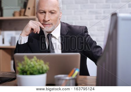 This looks interesting. Enthusiastic focused mature man looking at the screen of his laptop and carefully reading something while sitting at the desk in his office