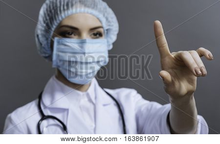 Look here. Female pretty concentrated doctor wearing hospital scrubs and gesturing while posing on grey background.