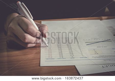Close up business man hand holding pen pointing stock market document in office on the wood desk.