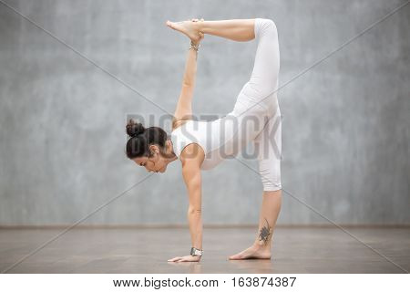 Profile portrait of beautiful young woman wearing white tank top working out against grey wall, doing yoga or pilates exercise. Standing in variation of Half Moon pose, Ardha Chandrasana. Full length