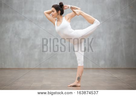 Profile portrait of beautiful young woman wearing white sportswear working out against grey wall, doing yoga or pilates exercise. Standing in Natarajasana, Lord of the Dance pose. Full length