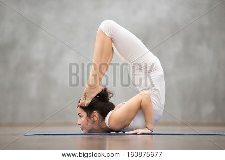 Side view portrait of beautiful young woman with tattoo on her foot meaning Wild kitty working out against grey wall, doing yoga or pilates exercise. Cheststand with backbend. Full length