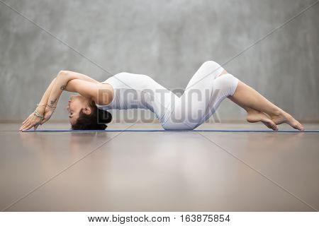 Beautiful young woman wearing white sportswear working out in fitness club against grey wall, doing yoga or pilates exercise. Variation of Matsyasana, Fish pose with crossed arms and legs. Full length
