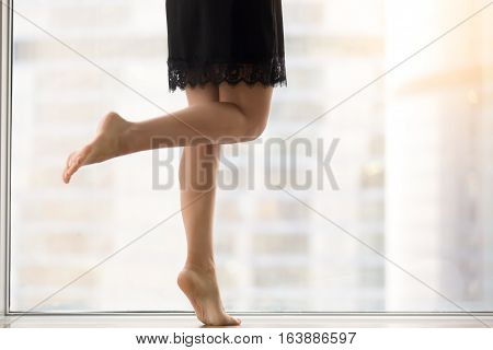Female bare legs, staying on tiptoe, healthy legs, smooth skin, helping mind and body relax and rejuvenate after work, taking dancing classes, exercising at morning at window with city view. Close up