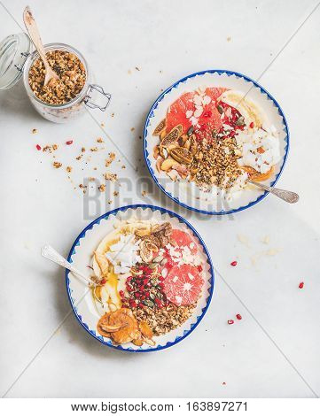 Healthy breakfast yogurt bowls with granola, grapefruit, banana, pomegranate, dried fruit, nuts, seeds over grey marble background, top view. Clean eating, detox, gluten-free, allergy-friendly concept
