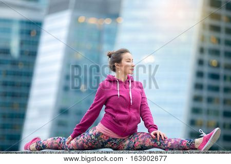 Young attractive woman practicing yoga, sitting in Monkey God exercise, Splits, Hanumanasana pose, working out wearing pink sportswear, outdoor full length, modern skyscraper background
