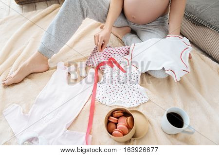 Home atmosphere. Close up of pregnant happy young woman posing with cute new baby clothes and food while reaxing and sitting in a cozy room.