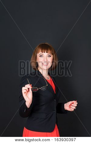 Cheerful confident business woman standing in dark office room and looking at camera holding glasses. Charming businesswoman on black background. Free space for text