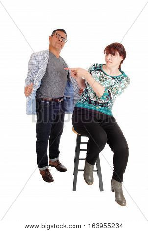 An wondering woman sitting on a chair pointing back at her standing husband looking pussled isolated for white background.