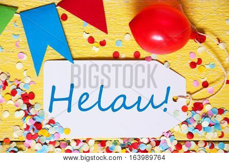 White Label With German Text Helau Means Happy Carnival. Close Up Of Party Decoration Like Streamer, Confetti And Balloon. Flat Lay Or Top View. Yellow Wooden Background