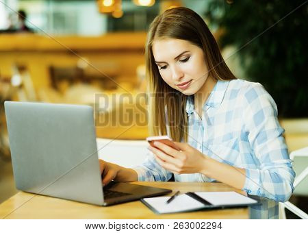 Young beautiful blond girl sitting in a coffee shop and working on computer. Image of happy woman using laptop and smartphone in cafe.