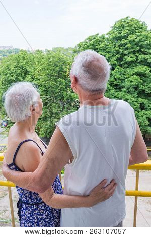 Senior Couple Standing Oustide At Balccony At Home And Looking Awayat Green Trees