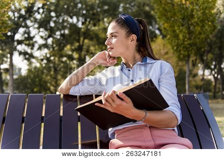 Beautiful  Young Woman  Holding  Open Book And Reading On Bench In Autumn Park
