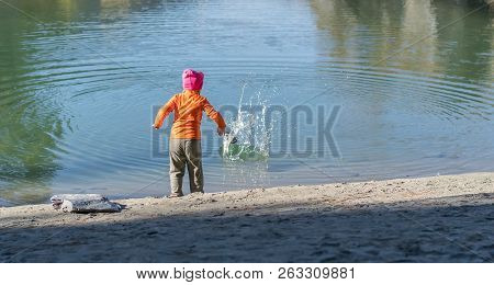 A Little Girl, A Child, In Warm Clothes And A Red Cap Throws Stones Into The Water From The Sandy Sh