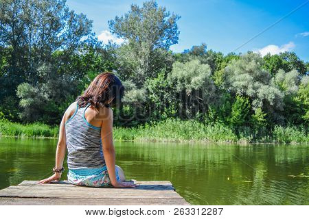 The Girl Sits On The River Dock, A Hot Summer Day, Rear View. Girl Sitting On Pier And Lookingat The