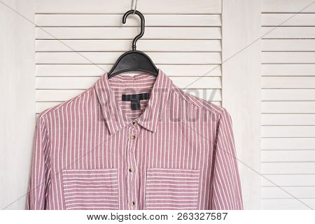 Pink Stried Shirt Hanging On A Hanger. White Wooden Screen On The Background. Fashionable Wardrobe