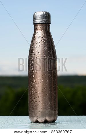 Thermo Stainless Bottle, Sprayed With Water. Sky And Forest On Background. On The Glass Desk. Thermo