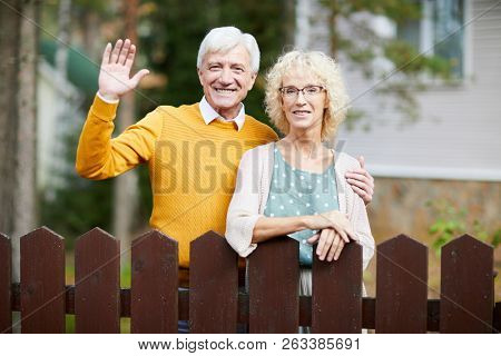 Hospitable mature couple standing by fence while friendly man waving hand to their neighbours