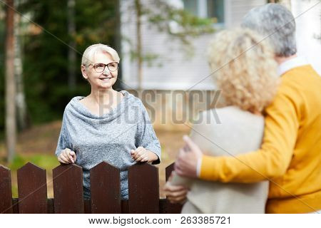 Blonde mature woman in eyeglasses and grey pullover looking at her neighbours during talk through fence