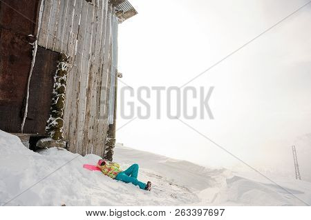 Girl Lying On The Snowboard Near The House In The Mountain And Looking Into A Distance In The Popula